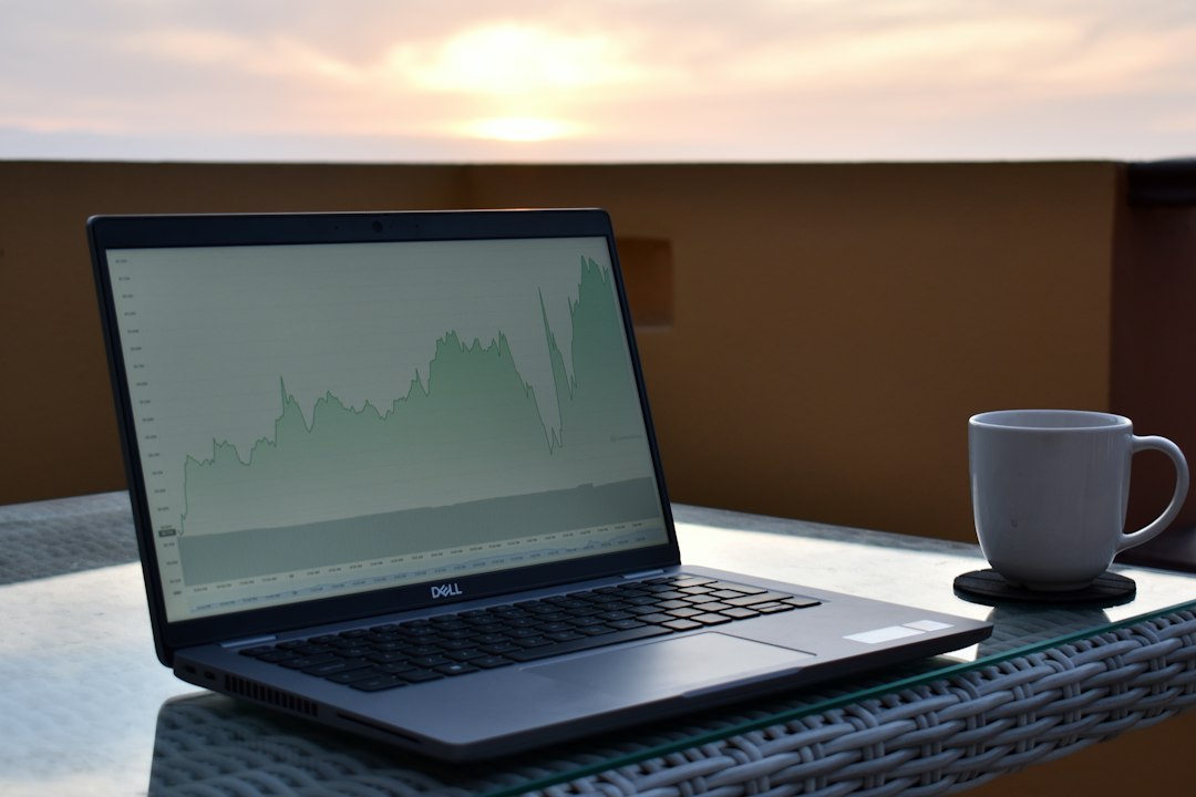 A laptop displaying a financial chart on a table with a cup of coffee, set against a sunset backdrop.